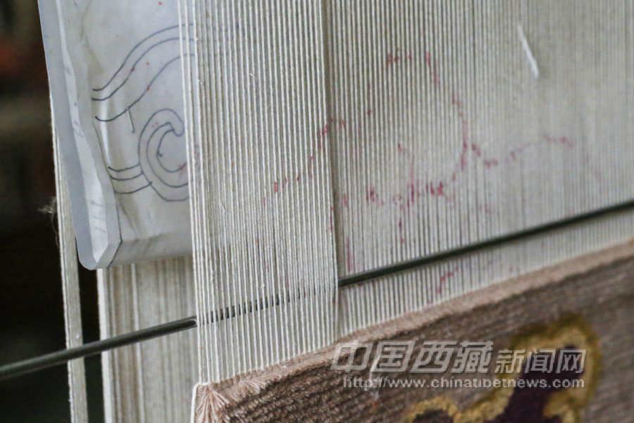將繪制好圖案的紙固定在經(jīng)線后方，然后用紅顏色的筆照著圖案的樣子描在經(jīng)線上。一切準(zhǔn)備就緒，開(kāi)始漫長(zhǎng)的藏毯編織過(guò)程。